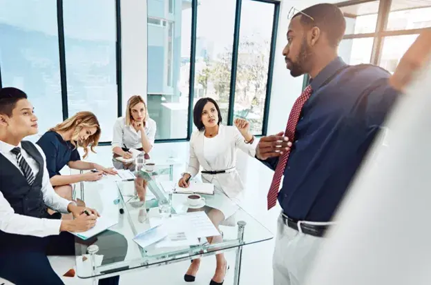 Business team in a modern office engaged in a discussion, with one person presenting and others listening attentively, representing the importance of addressing all three agendas of leadership for holistic, effective leadership.